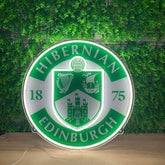 A circular illuminated Hibernian Edinburgh sign with a green and white design, displayed on a wooden surface against a green ivy wall background.