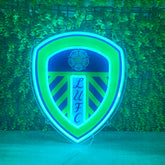 A shield-shaped LED neon sign displaying the Luton United Football Club crest, glowing with blue and green light against a green ivy-covered wall.