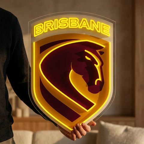 A person holds a large shield-shaped Brisbane sports sign with a glowing yellow 'BRISBANE' top and a dark red horse emblem outlined in yellow, shown in a softly lit indoor setting.