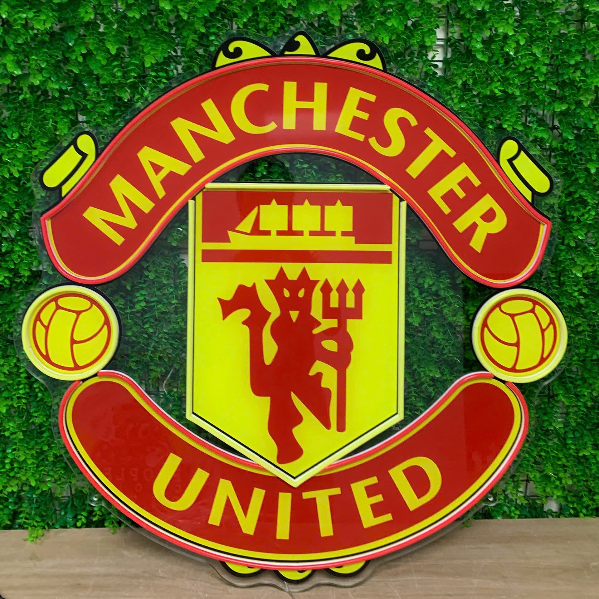 A large Manchester United crest wall sign with red and yellow colors, displayed against a green ivy wall backdrop.