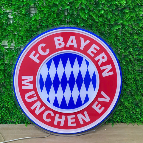 A large circular FC Bayern München EV sign featuring the club's red, white, and blue logo, displayed against a green ivy-covered wall.
