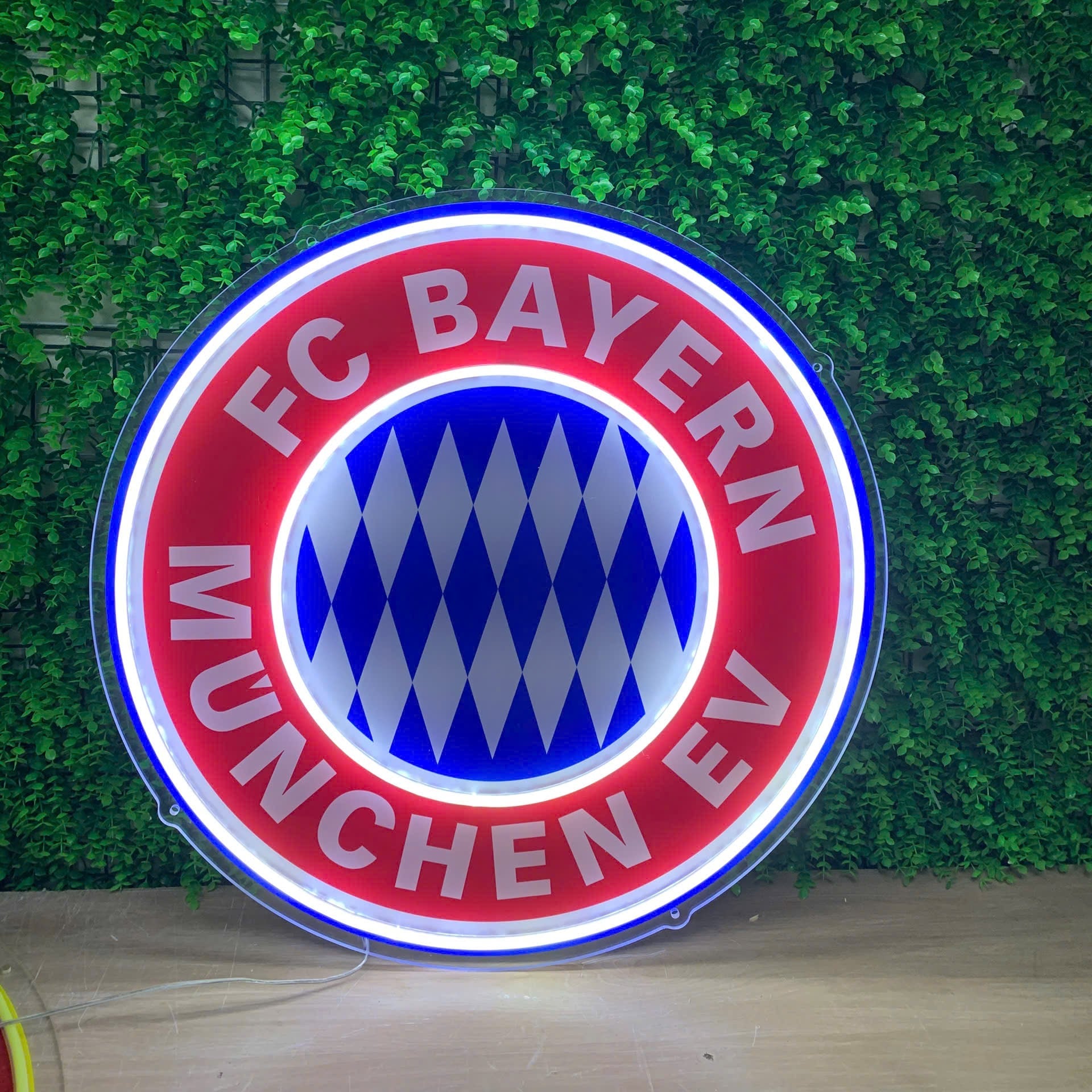 A circular illuminated FC Bayern Munich LED neon sign displayed against a green artificial ivy wall backdrop.
