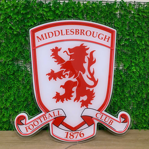A Middlesbrough Football Club crest sign featuring a red lion rampant on a white shield, with 'MIDDLESBROUGH', 'FOOTBALL CLUB', and '1876' text, displayed against a green hedge wall.