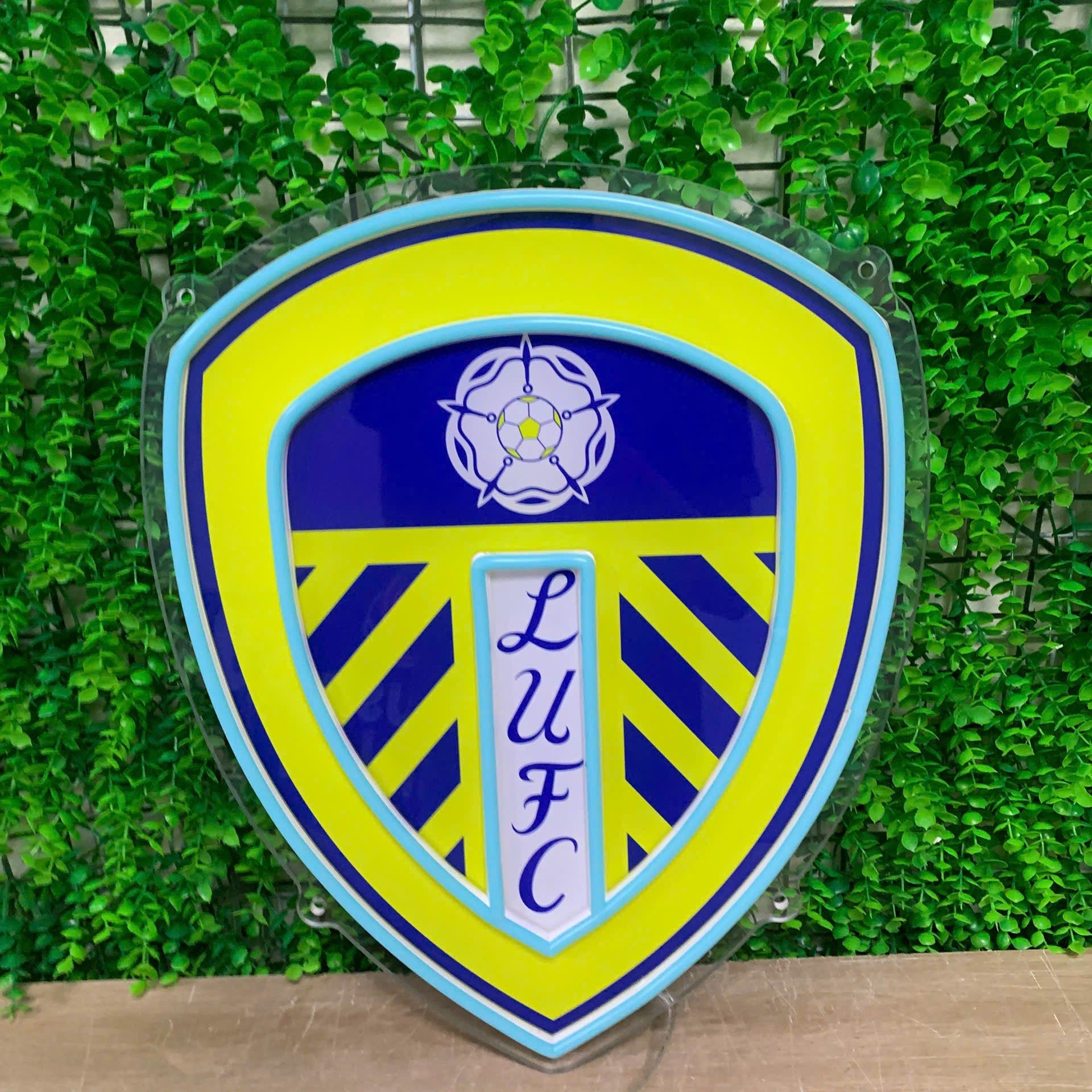 A shield-shaped LED sign featuring the Leeds United Football Club crest, with a bright yellow and blue design, mounted against a green artificial foliage wall.