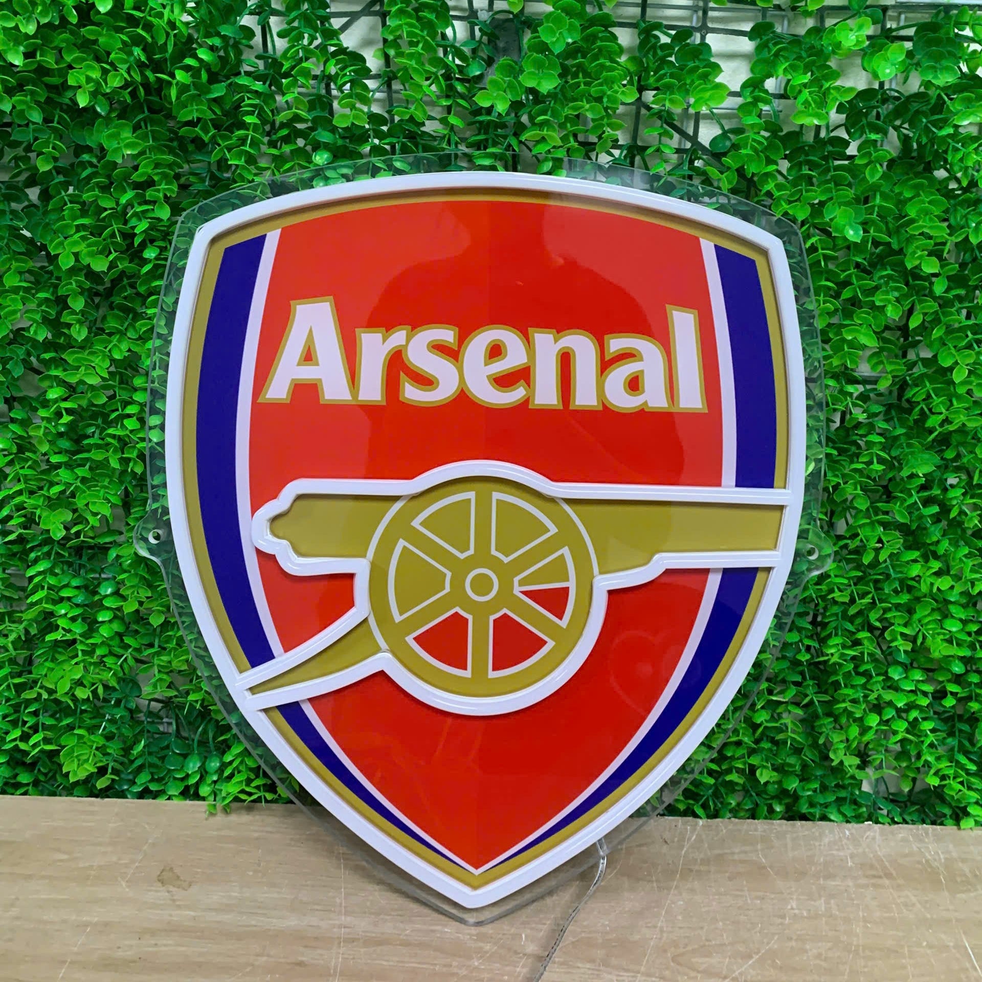 An Arsenal football club crest sign with a glossy red, white, blue, and gold design, displayed against a green artificial ivy wall.