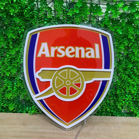 An Arsenal football club crest sign with a glossy red, white, blue, and gold design, displayed against a green artificial ivy wall.