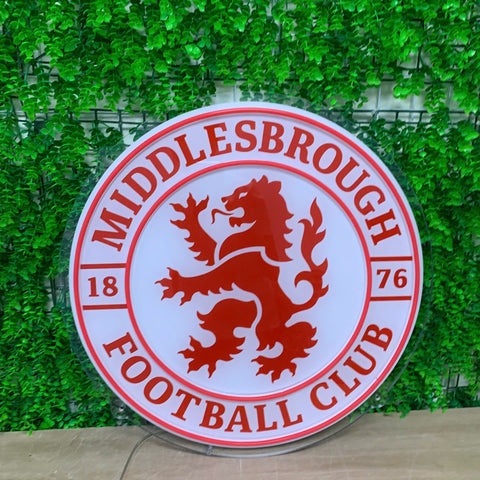 A circular Middlesbrough Football Club sign featuring the club's red lion crest on a white background, with 'MIDDLESBROUGH FOOTBALL CLUB' and the years '1876' printed in red. The sign is displayed on a wooden surface in front of a green artificial ivy wall.