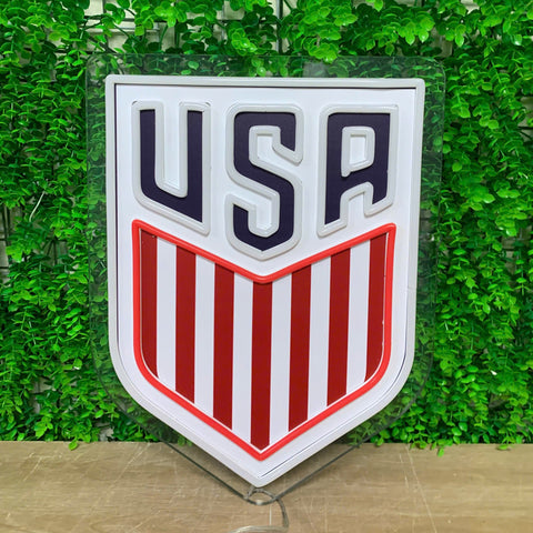 A shield-shaped USA soccer crest sign with red and white vertical stripes and dark navy lettering, mounted on a clear acrylic stand against a green leafy wall backdrop.
