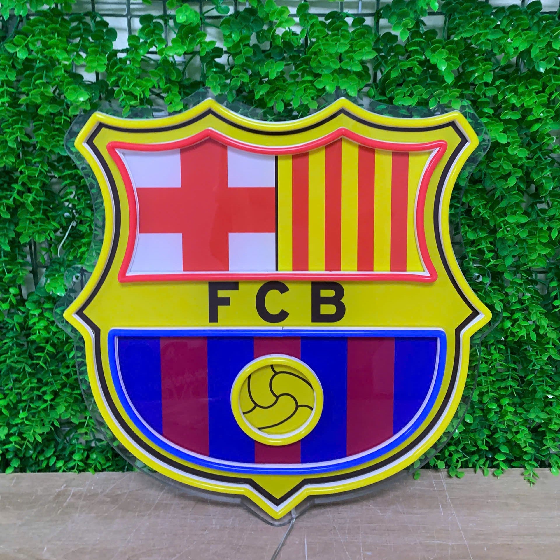 A large FC Barcelona shield crest sign with yellow, red, blue, and white sections, mounted against a green leaf wall.
